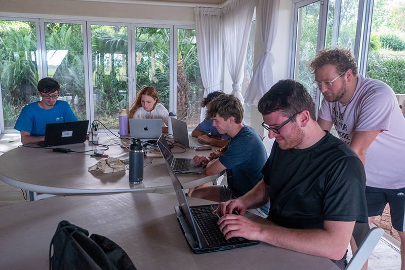 Group of people working on laptops around a large table in a bright room with floor-to-ceiling windows overlooking greenery.