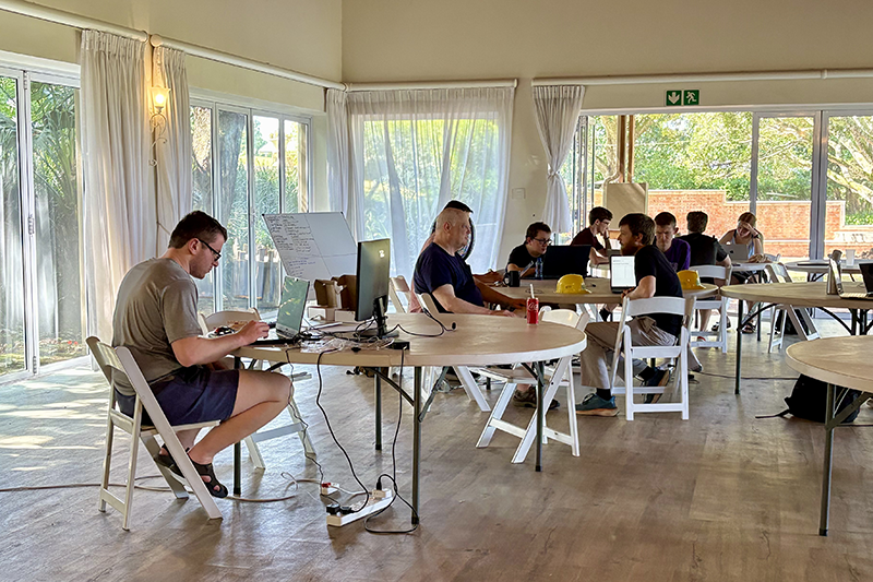Small group of people working on laptops at shared tables in a sunlit room with floor-to-ceiling windows and white curtains.
