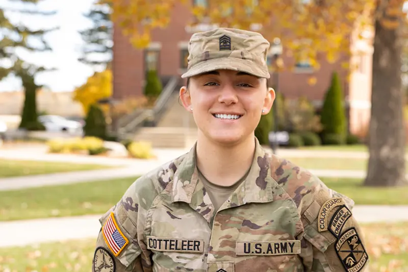 Isabel Cotteleer ROTC cadet in camouflage uniform smiling outdoors, with a campus building and autumn foliage in the background.