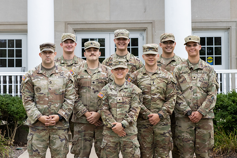 Group of rotc cadets in camouflage uniforms posing together in front of a building with white columns.