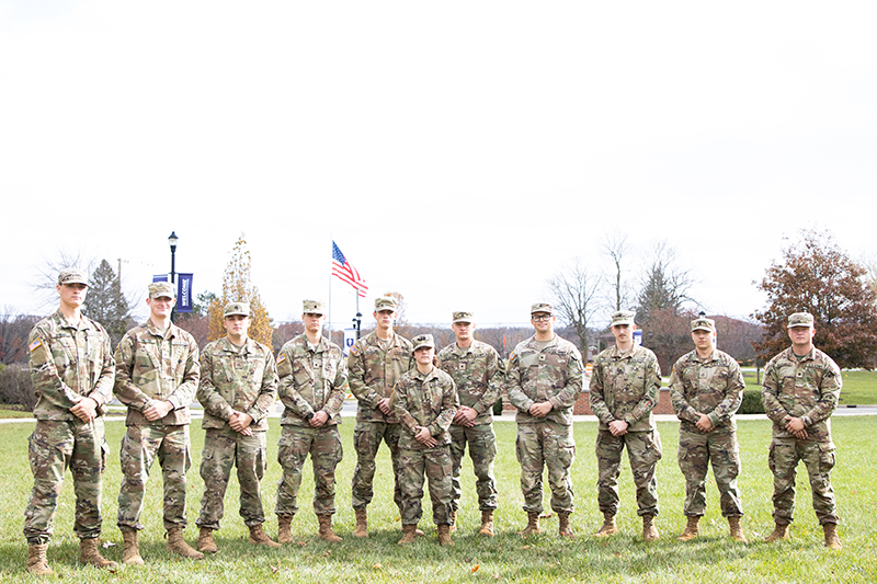 Group of rotc cadets in camouflage uniforms standing in a line on a grassy field, with an American flag in the background.
