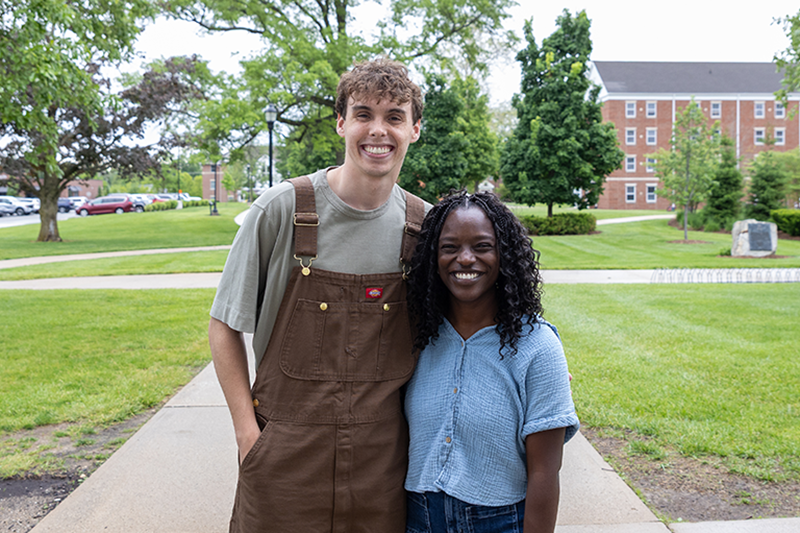 Ethan and Hasi smiling with arms around each other on a college campus walkway, with trees and a brick building in the background.