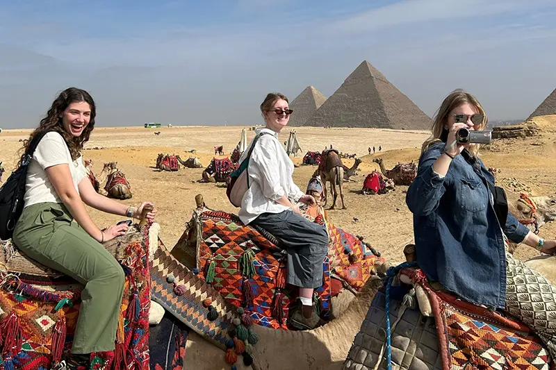 Three girls on camels with Egyptian pyramids in background.