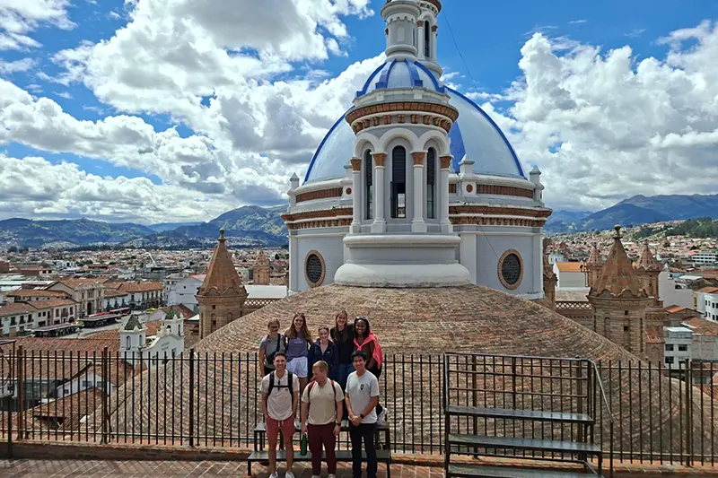 Group of people posing on a terrace overlooking a city, with a large blue-and-white domed cathedral and mountains under a cloudy sky in the background.