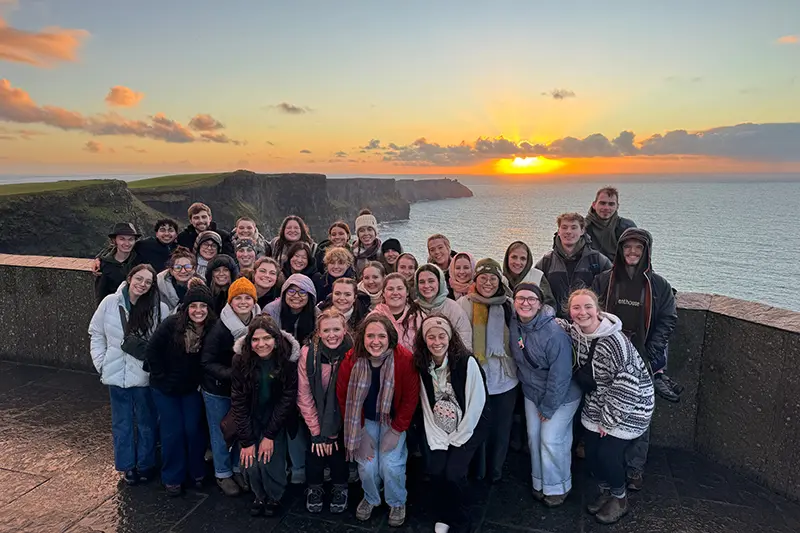 Large group of people posing together at a cliffside overlook by the ocean at sunset, with sea cliffs and clouds in the background.