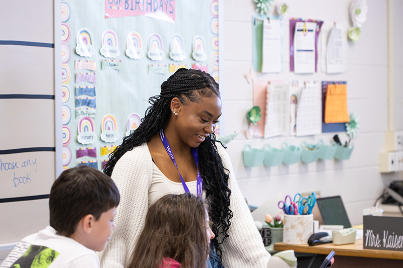 Student teacher in a classroom smiles while helping two young students at a desk, with colorful posters and supplies in the background.