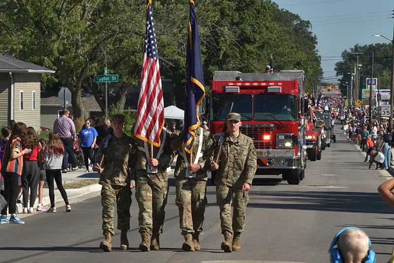An ROTC honor guard carrying the American flag and state flags, marching in a parade with a fire truck in the background and a crowd of spectators lining the street.