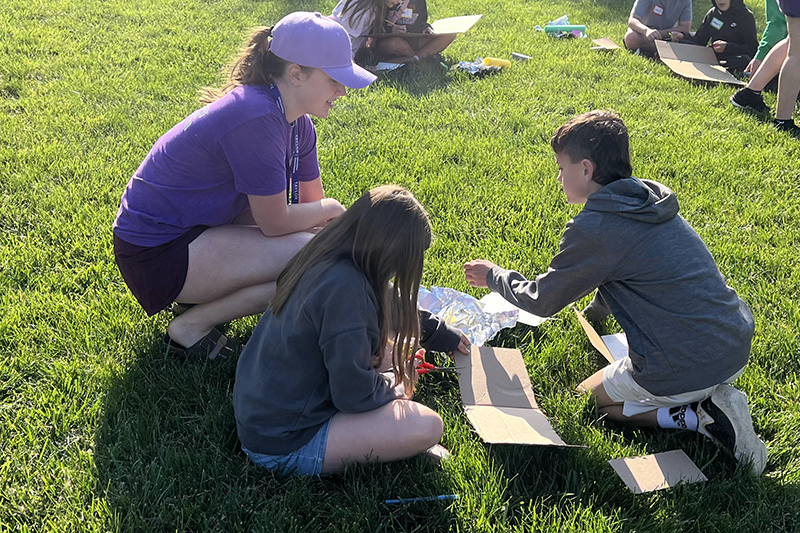 Taylor student kneels on the grass, helping two kids assemble a cardboard project during an outdoor activity.