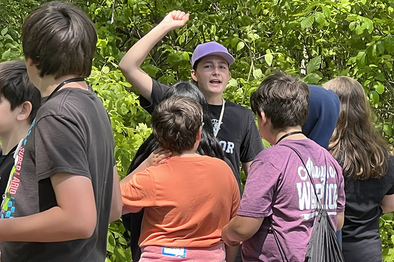 Group of kids outdoors in a wooded area, with a Taylor student in a purple cap raising an arm while talking to the others.