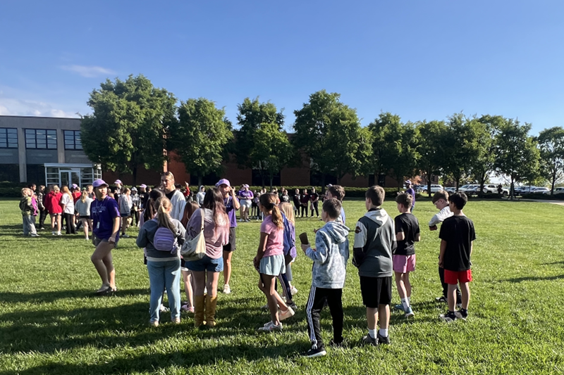 Students gather on a school lawn for an outdoor activity, with lines of participants stretching across the grass under a clear blue sky.