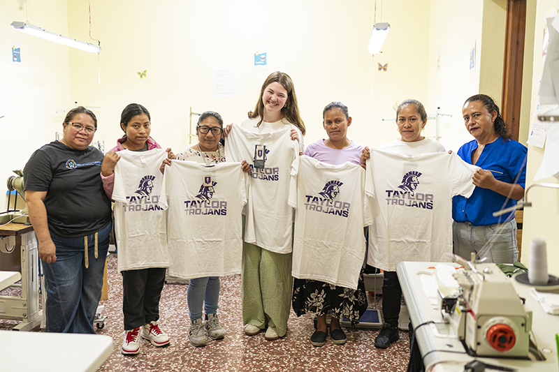 Group of women in a sewing workshop holding up matching white "Taylor Trojans" T-shirts, with sewing machines visible in the room.