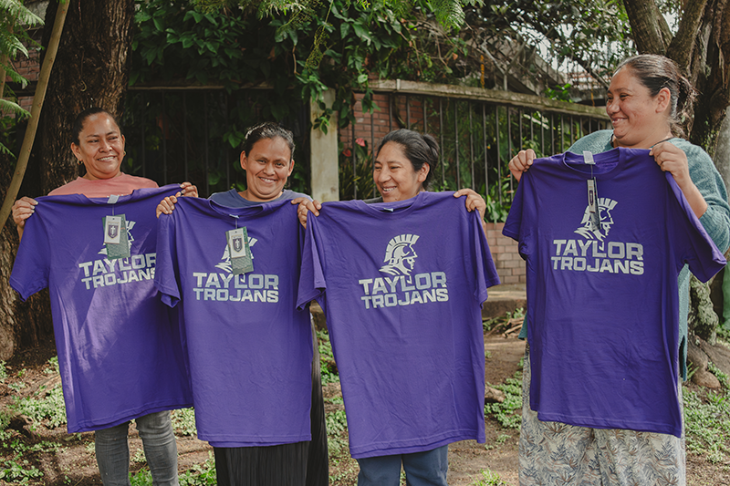 Four women outdoors holding up purple "Taylor Trojans" T-shirts and smiling.