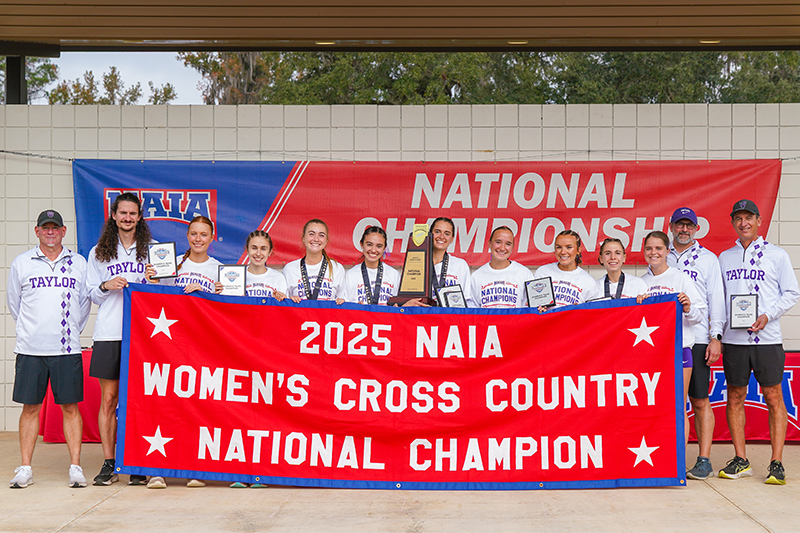 Women’s cross country team and coaches pose on a stage holding awards behind a large banner reading “2025 NAIA Women’s Cross Country National Champion.”