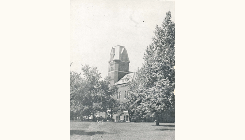 Historic black-and-white photo of a brick building with a tall tower, partly obscured by trees across a grassy lawn.