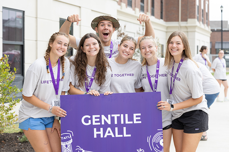 Group of smiling students in matching T-shirts and lanyards pose outdoors behind a large purple “Gentile Hall” sign.